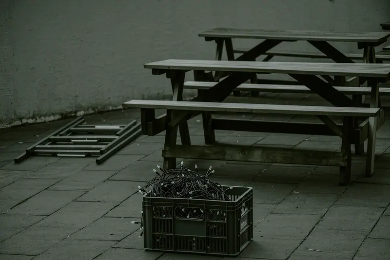Stacked bench sets, a folded tent frame and a crate of festoon lighting standing on paving in a courtyard.