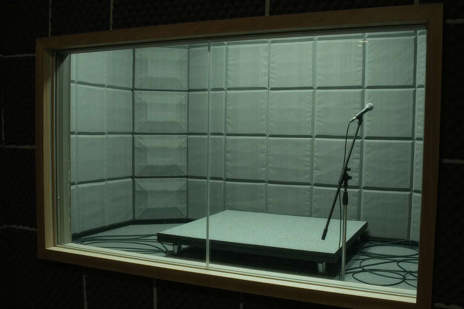 A small rehearsal room with grey acoustic foam panels on the walls and a bare carpeted drum riser, coiled cables and an empty mic stand on the floor in the foreground.