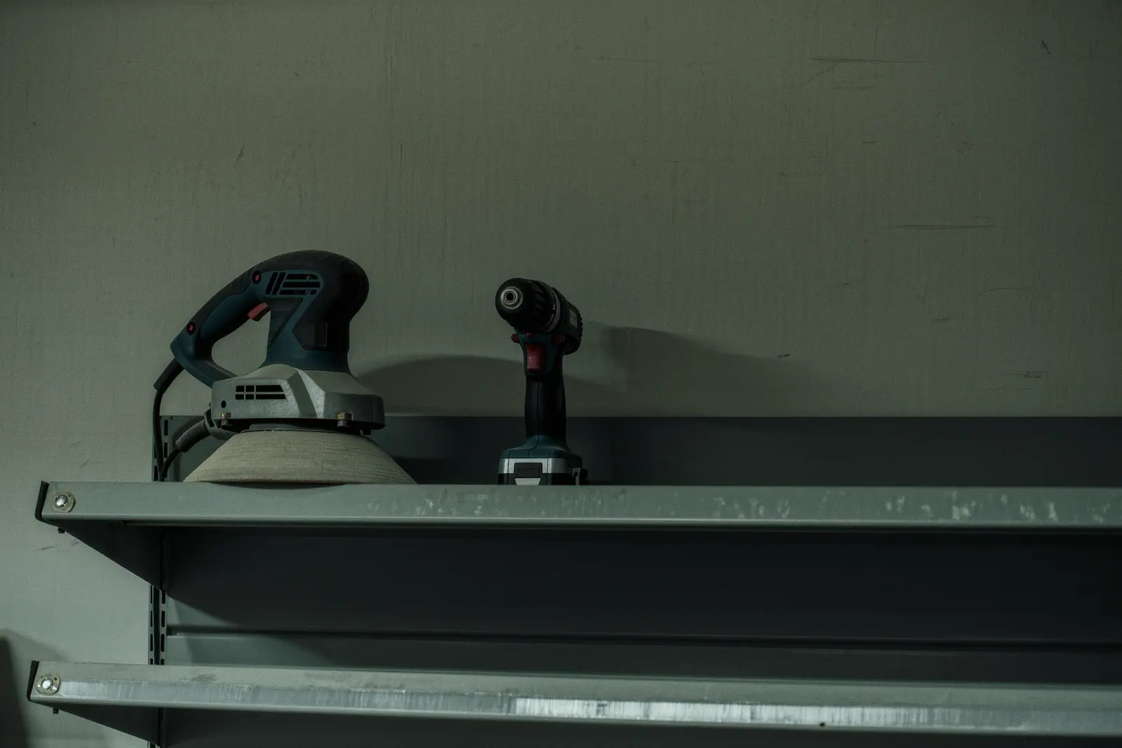 Hire power tools and a stripped floor sander on a steel shelving unit in a plain store room, the sander's drum and bent handle close in the foreground.