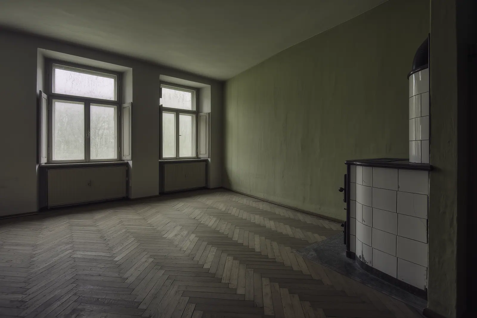 An empty Altbau room with herringbone parquet, a tall tiled stove in the corner and bare double casement windows, the worn boards running into the foreground.