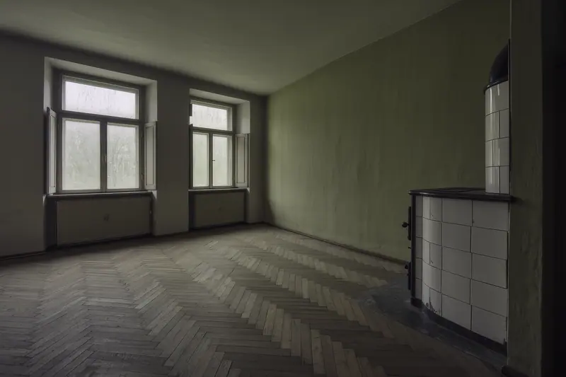 An empty Altbau room with herringbone parquet, a tall tiled stove in the corner and bare double windows, cleared ready for a move.