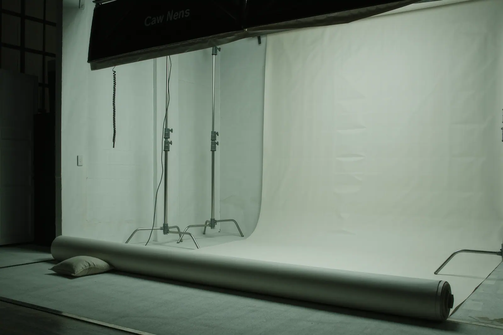 An empty white photo studio with a paper backdrop roll swept down onto the floor, two bare light stands and a ceiling power drop, a sandbag on the floor in the foreground.