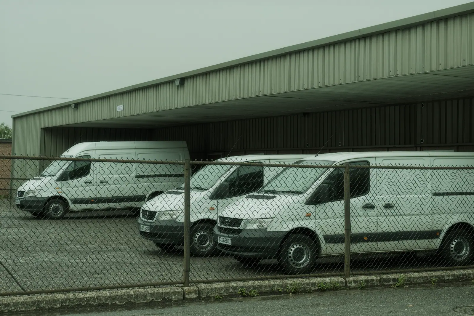 A small van hire depot yard with three plain white vans parked nose-in on cracked asphalt, a chain-link fence and its concrete footing across the foreground.