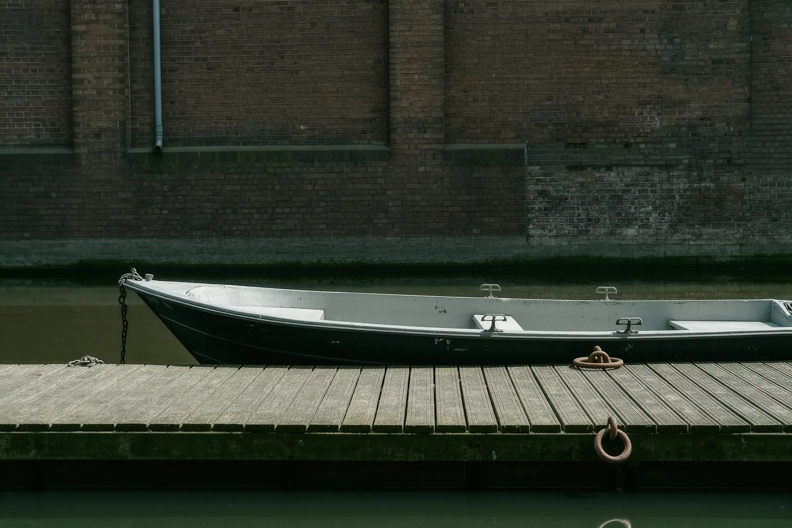 A small licence-free motorboat at a wooden jetty on a Berlin canal, weathered planks and a rusted mooring ring in the foreground, dark brick warehouse walls behind.