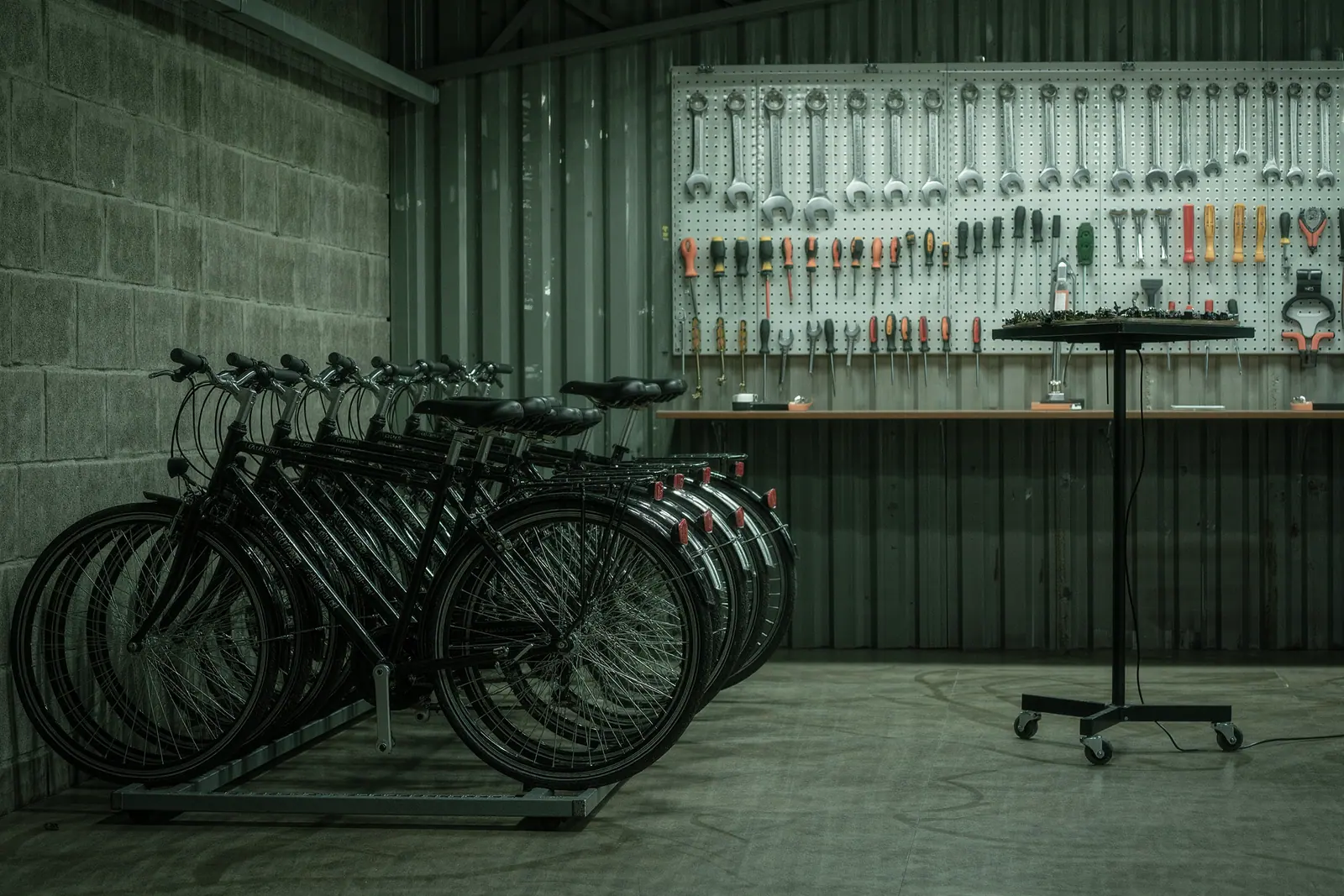 A rental bike shop interior with plain city bikes packed into a floor rack and a pegboard of tools on the wall, a repair stand and a tray of small parts in the foreground.
