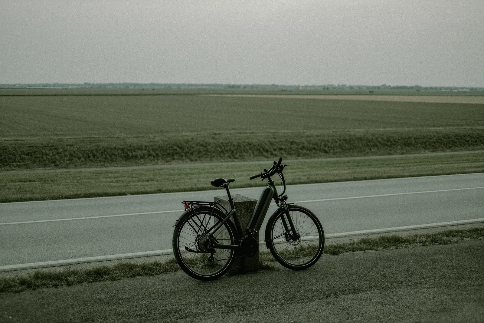 An e-bike leaning against a low concrete bollard beside a wide asphalt cycle path through flat open grassland at the city edge, the bollard and front wheel in the foreground.
